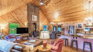 Interior acolhedor de uma cabana com sala de estar, cozinha e sala de jantar, apresentando paredes e teto de madeira, lareira e televisão.