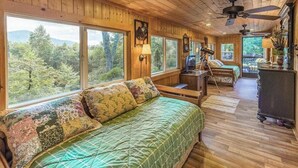 Uma cabana com uma cama grande, uma escrivaninha e uma cadeira. Várias janelas oferecem vista para a floresta.