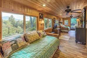 Eine Hütte mit einem großen Bett, einem Schreibtisch und einem Sessel. Mehrere Fenster bieten Blick auf den Wald.