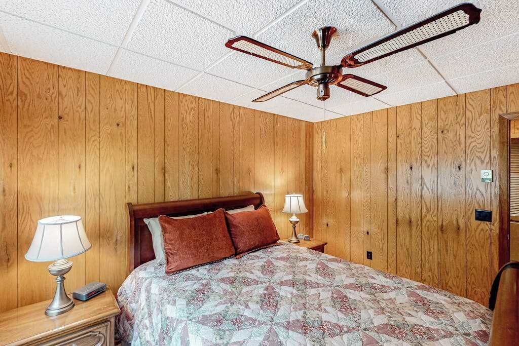 Un dormitorio con paneles de madera, una cama con colcha estampada, dos lámparas de noche, una mesita de noche y un ventilador de techo.