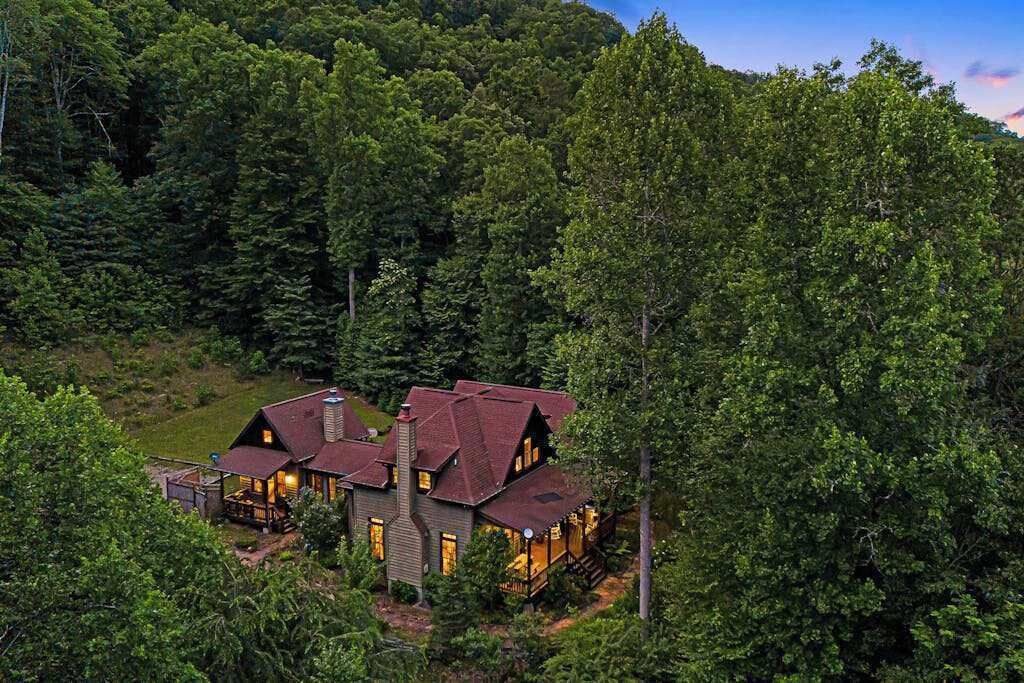 A large, two-story house surrounded by dense forest, with a prominent chimney and multiple windows.