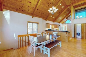 A spacious dining area with a wooden table and chairs, a kitchen in the background, and a high ceiling with wooden beams. - Mountain Hideaway (Almond)