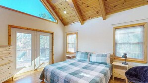 Ein Schlafzimmer mit einem großen Bett, Holzbalkendecke, Kommode, Stuhl und einem Fenster mit Jalousien.