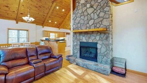 Ein Wohnzimmer mit Kamin aus Stein, einer Ledersofa und Holzboden, im Hintergrund eine Küche.