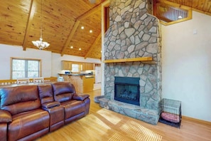 A living room with a stone fireplace, leather sofa, wooden ceiling, and a kitchen in the background. - Mountain Hideaway (Almond)
