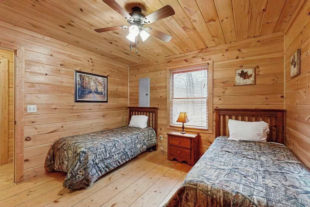A wooden cabin bedroom with two beds, a ceiling fan, a window with blinds, and a framed picture on the wall.
