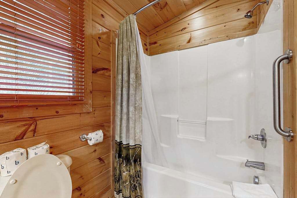Une salle de bain rustique, située dans une cabane en bois, équipée d’une douche, de toilettes et de stores aux fenêtres.