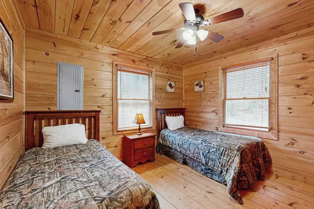 A wooden cabin bedroom with two beds, a ceiling fan, and windows with blinds.