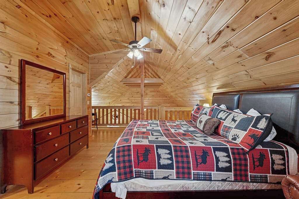 A wooden cabin bedroom with a bed, dresser, ceiling fan, and a large mirror.