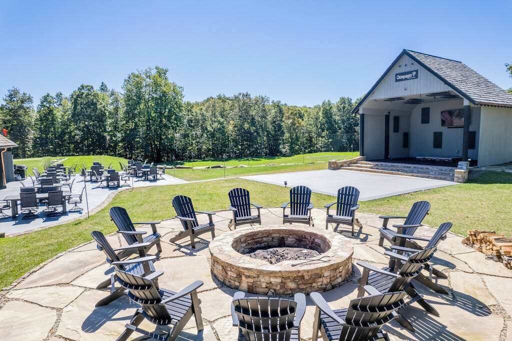 A patio with Adirondack chairs, a stone fire pit, and a building labeled 'Chimney House' in the background.