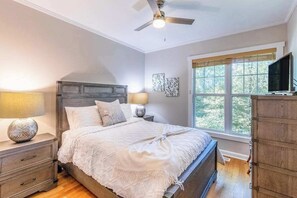 A bedroom with a bed, nightstands, a ceiling fan, a window with blinds, and a television on a stand. - Carolina Cottage (Highlands)