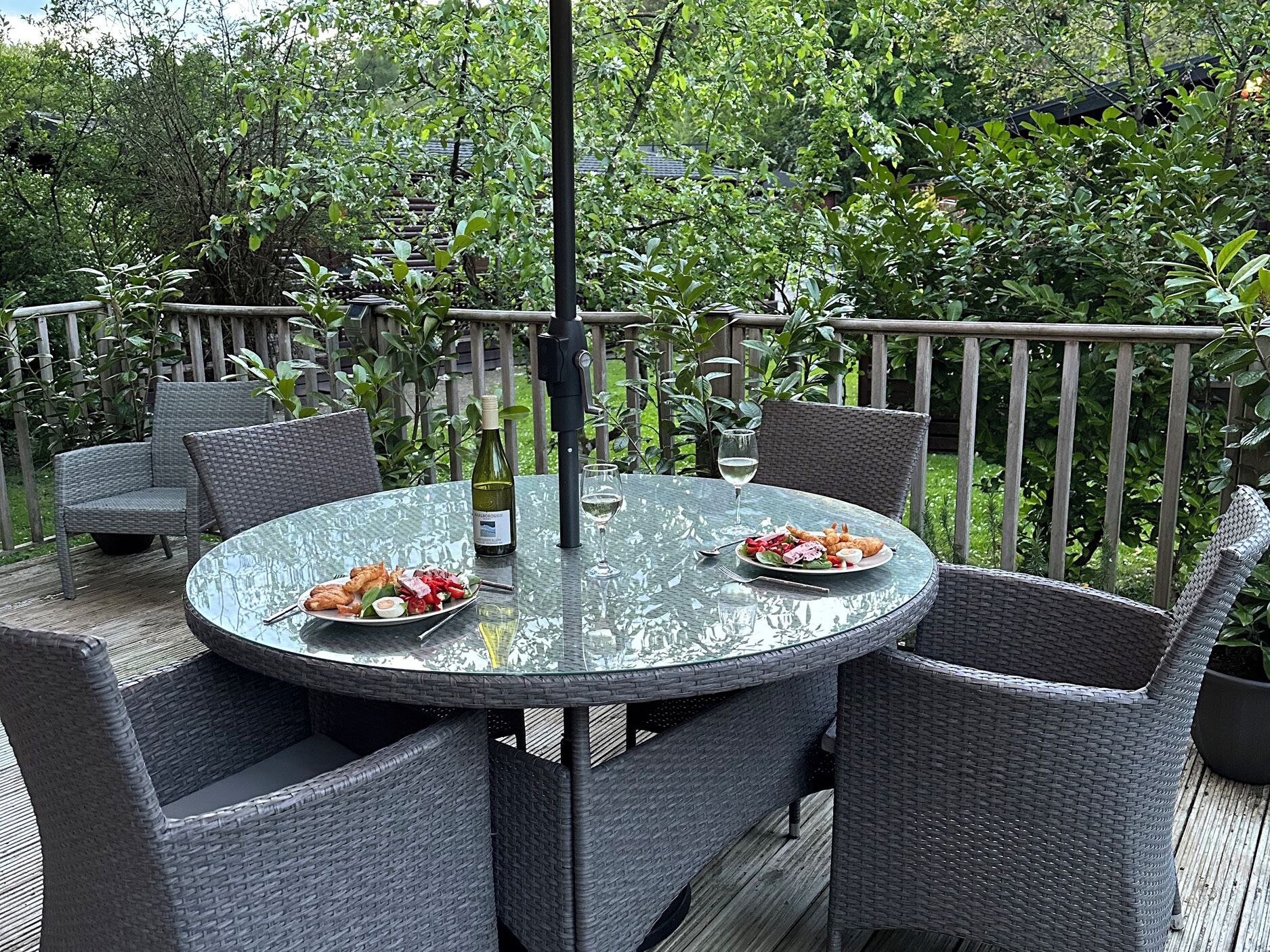 Een ronde glazen tafel met rieten stoelen, een fles wijn en een glas witte wijn op een terras met houten leuning en weelderige begroeiing op de achtergrond.