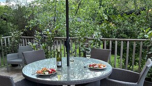 A round glass table with wicker chairs, a bottle of wine, and a glass of white wine on a deck with a wooden railing and lush greenery in the background. - Tiggywinkle Beck - White Cross Bay (Windermere)