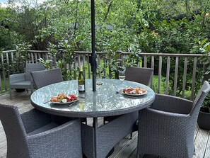 Ein runder Glastisch mit Korbstühlen, eine Weinflasche und ein Glas Weißwein auf einer Terrasse mit Holzwertung und üppigem Grün im Hintergrund.