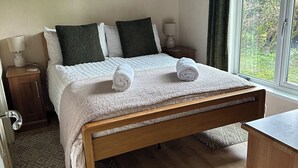 A neatly made bed with a white duvet and rolled towels, a wooden bench, a nightstand with a lamp, and a window with a view of greenery. - Tiggywinkle Beck - White Cross Bay (Windermere)