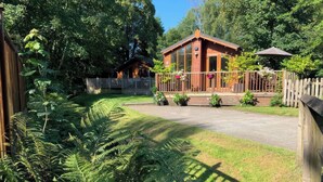 A wooden cabin with a porch, surrounded by lush greenery and a wooden fence. - Tiggywinkle Beck - White Cross Bay (Windermere)