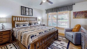 Un dormitorio con una cama de madera, un ventilador de techo, una ventana con persianas, un sillón gris y una alfombra.