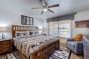 A bedroom with a wooden bed, a ceiling fan, a window with blinds, a gray armchair, and a rug. - The Mountain Haven (Maggie Valley)
