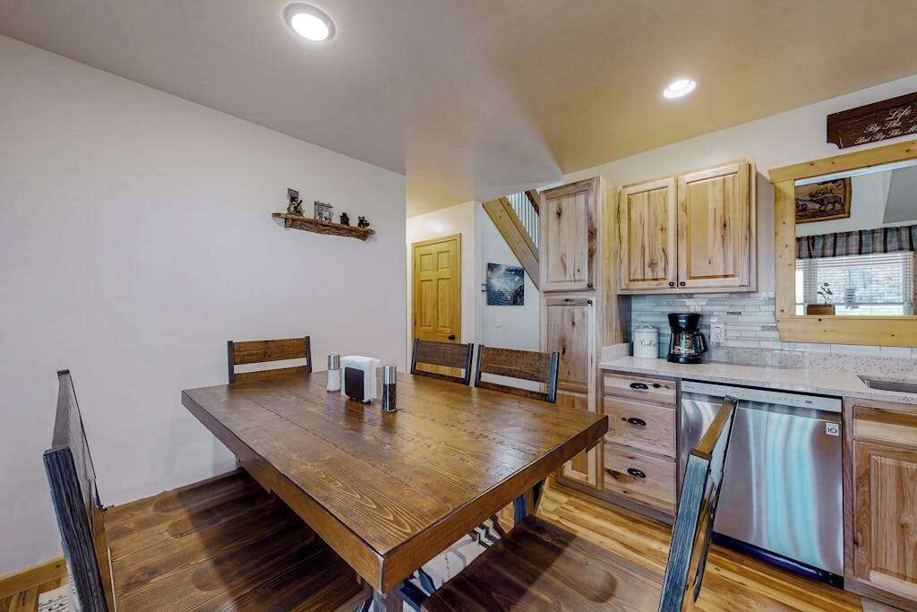 A dining area with a wooden table and chairs, a kitchen with wooden cabinets and a stainless steel dishwasher, and a staircase leading to another level.