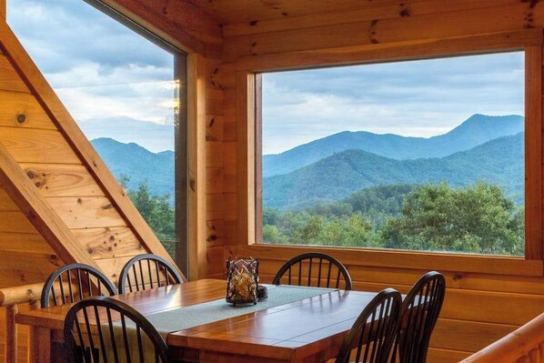 Ein Holzinnenraum mit einem Esstisch und Stühlen, ein großes Fenster mit Blick auf die Berge.