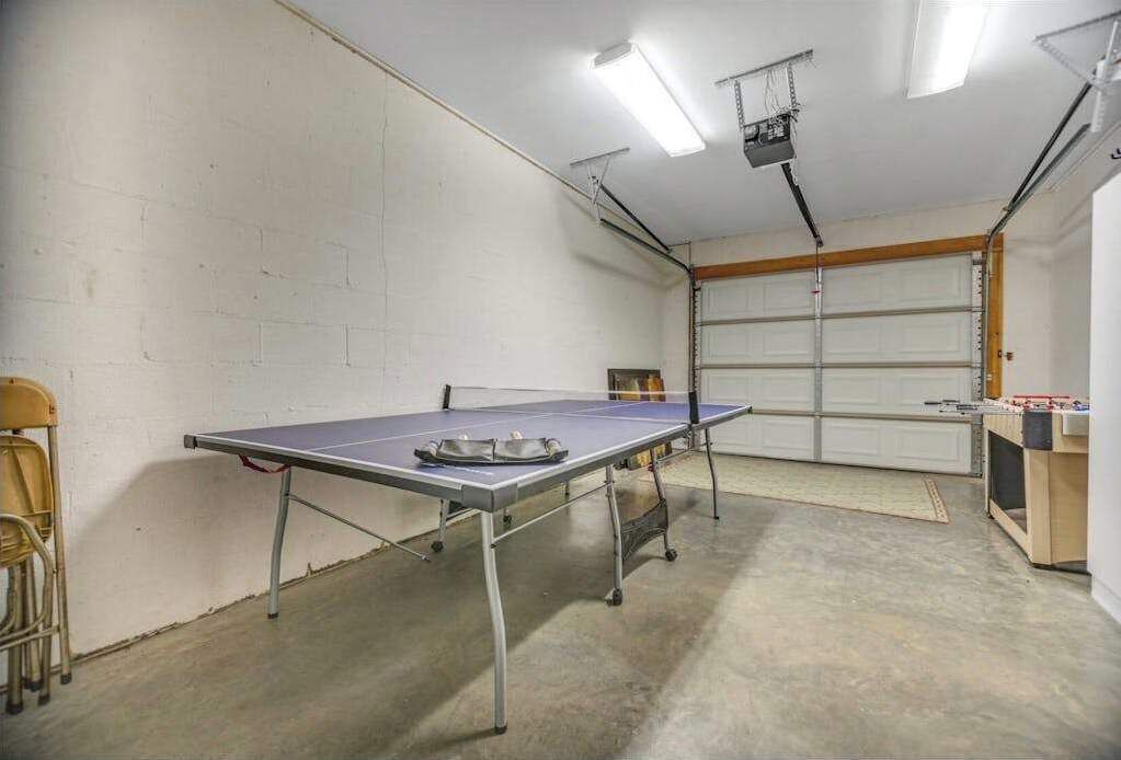 A ping pong table in a garage with a white wall, a wooden door, and fluorescent lighting.