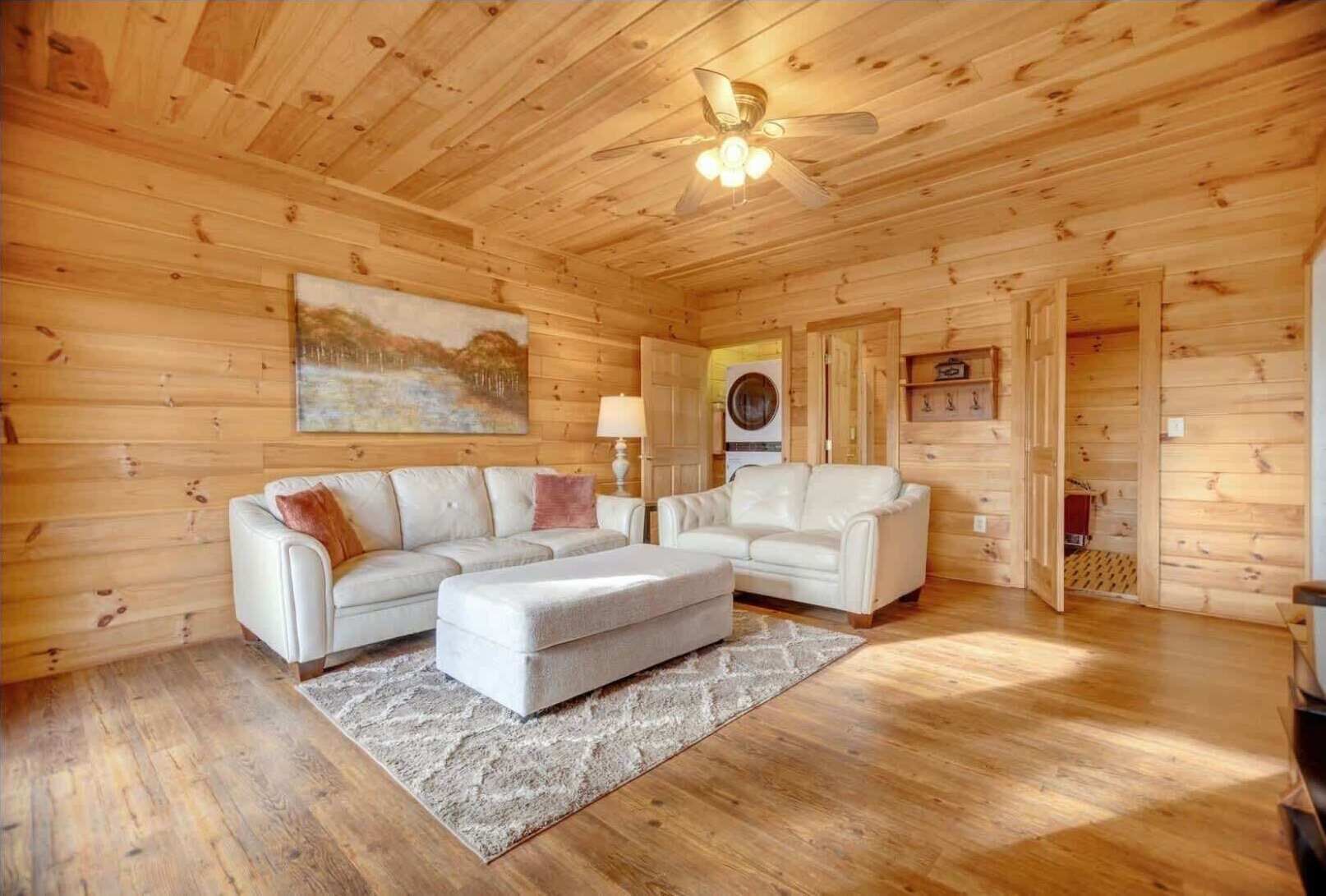 A wooden cabin interior with a sofa, ottoman, ceiling fan, and a painting on the wall.