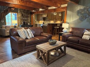 Ein gemütliches Wohnzimmer mit zwei braunen Ledersofas, einem Holztisch und einer Küchenzeile mit Steinwänden und Holzbalken.