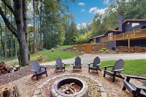 Eine Hütte mit Feuerstelle, umgeben von Adirondack-Stühlen, einer Terrasse und einem bewaldeten Hintergrund.