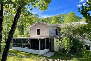 Ein Haus mit einem großen Glasanbau, umgeben von Bäumen und einer Hügelkulisse im Hintergrund.