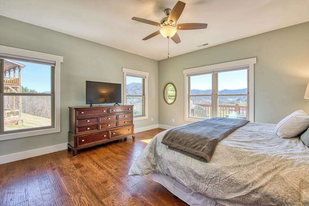 A bedroom with a bed, dresser, television, ceiling fan, and large windows.