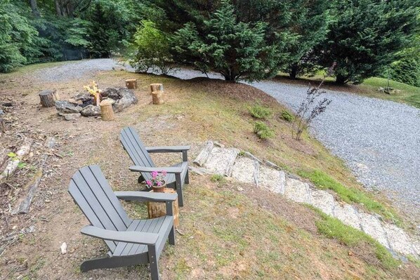Un terrain de camping équipé de deux fauteuils Adirondack, d’un foyer et d’un chemin de gravier menant à une zone boisée.