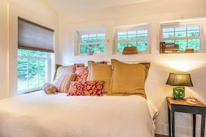A bedroom with a bed, pillows, a bedside table with a lamp, and windows with views of greenery. - Fabulous escape with firepit, stylish interior & central AC - near hiking (Waynesville)