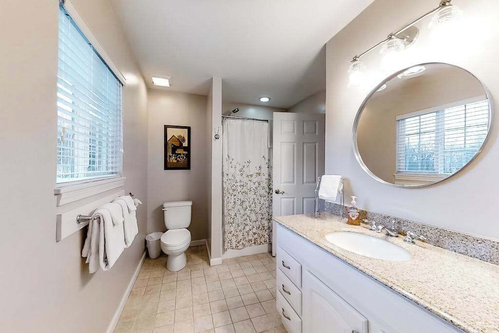 Un baño con inodoro, ducha con cortina estampada, lavamanos con espejo y ventana con persianas.