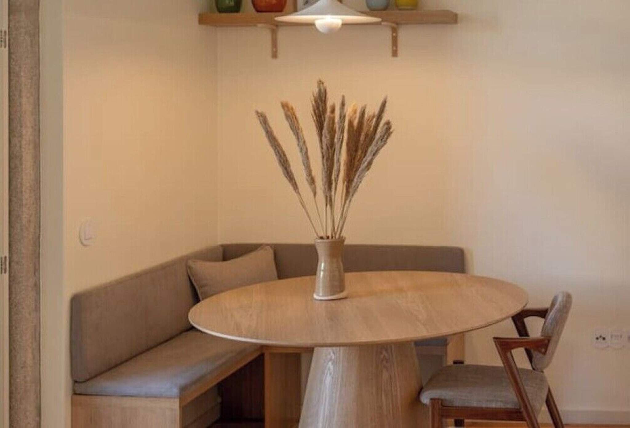 Een ronde houten tafel met een vaas droogbloemen, een grijze bank en een houten stoel in een kamer met een lamp erboven.