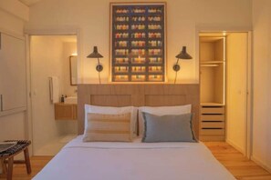 Quarto com cama, luminárias de cabeceira, cabeceira, prateleira na parede com garrafas, armário e banheiro.
