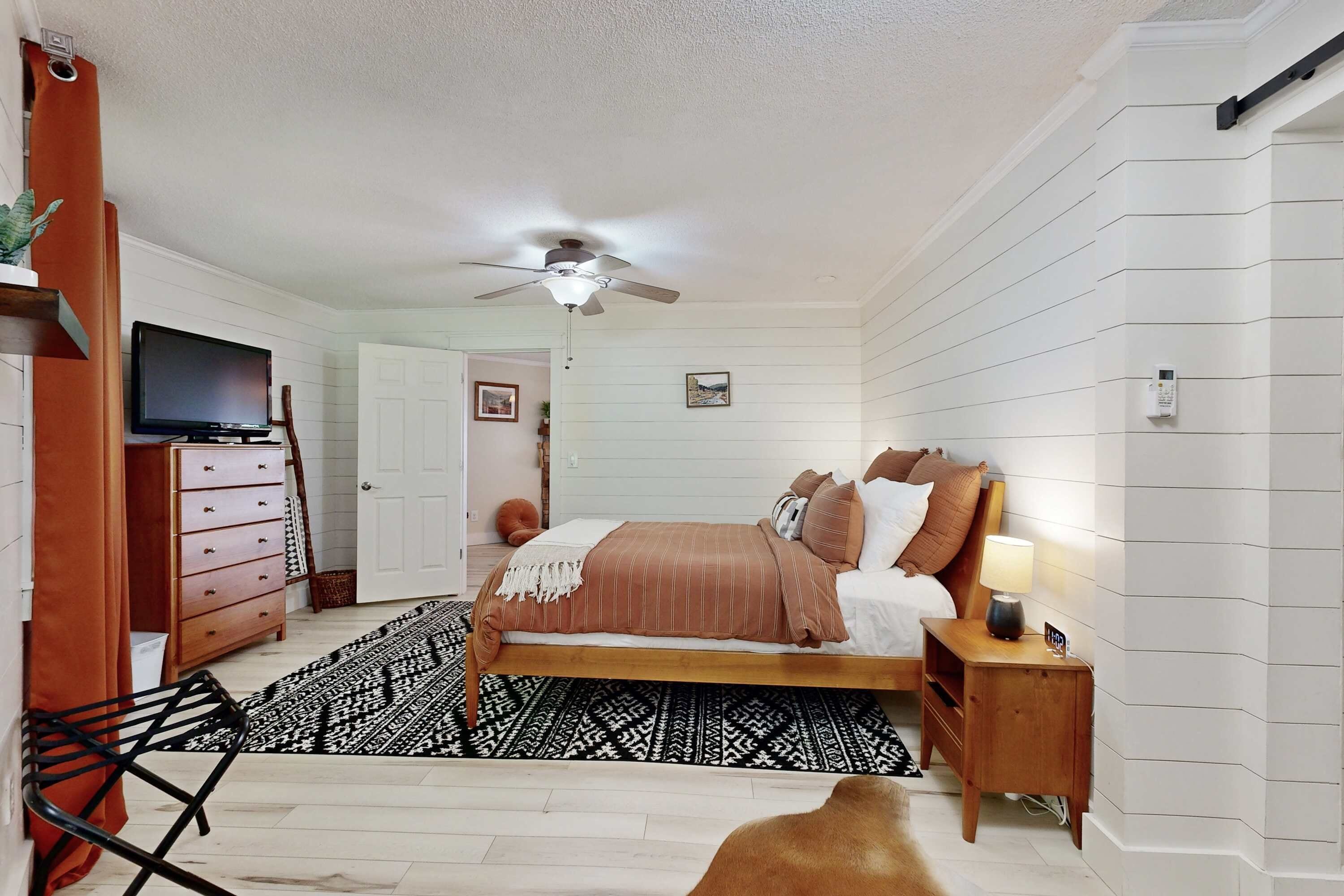 A bedroom with a bed, a ceiling fan, a TV, a dresser, and a nightstand with a lamp.