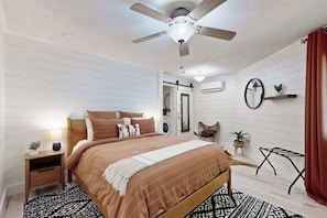 Ein Schlafzimmer mit Bett, Deckenventilator, Nachttisch, Sessel, Spiegel, Regal mit Pflanze und Fenster mit Vorhängen.
