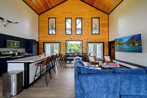 A spacious interior with a kitchen, dining area, and living room, featuring wooden walls, large windows, and a central island. - New! Heated Pool, Riverfront, Kayaks, Pool Table (Broken Bow)