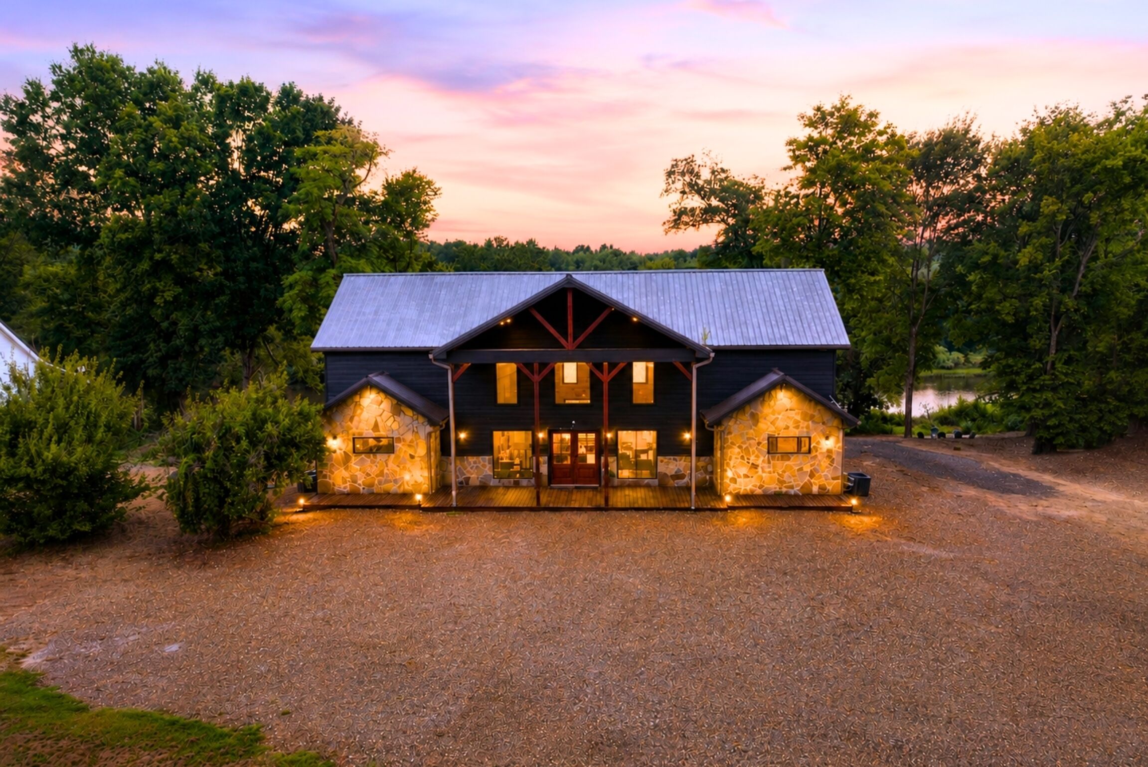 A two-story building with a stone facade and a metal roof, surrounded by trees and a gravel driveway.