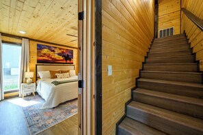 A wooden interior with a bed, nightstand, and a painting of a sunset. - New! Heated Pool, Riverfront, Kayaks, Pool Table (Broken Bow)