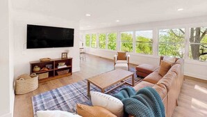 Ein geräumiges Wohnzimmer mit großem Fernseher, bequemen Sitzgelegenheiten und Blick durch große Fenster.