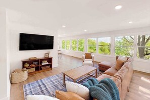 A spacious living room with a large TV, comfortable seating, and a view through large windows. - Inviting home with enclosed yard, mountain view, firepit, & W/D (Waynesville)