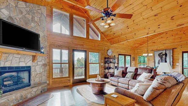 A log cabin interior with a stone fireplace, leather sofa, wooden floors, and a ceiling fan. - The Glens of Iron Duff (Waynesville)