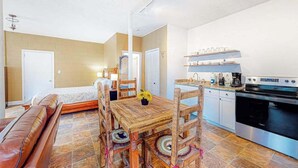 Een gezellige kamer met een houten eettafel, stoelen, een bank, een keukenhoek met een fornuis en kastjes, en een deur naar een andere kamer.