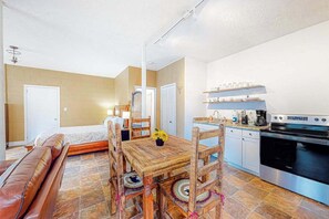 A cozy room with a wooden dining table, chairs, a sofa, a kitchen area with a stove and cabinets, and a door leading to another room. - Cozy Mountain Hideaway (Waynesville)