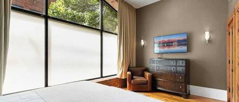 A bedroom with a large window, a ceiling fan, a bed, a chair, a TV, and a wooden dresser.