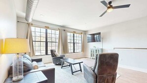 Ein modernes Wohnzimmer mit einer Couch, Sesseln, einem Couchtisch und einer Lampe. Große Fenster mit Vorhängen, ein Deckenventilator und ein flachbildfernseher an der Wand sind ebenfalls vorhanden.