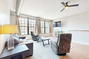 A modern living room with a sofa, armchairs, a coffee table, and a lamp. There are large windows with curtains, a ceiling fan, and a flat-screen TV mounted on the wall. - Spacious, chic retreat with balcony & AC - walk to dining & shopping (Asheville)