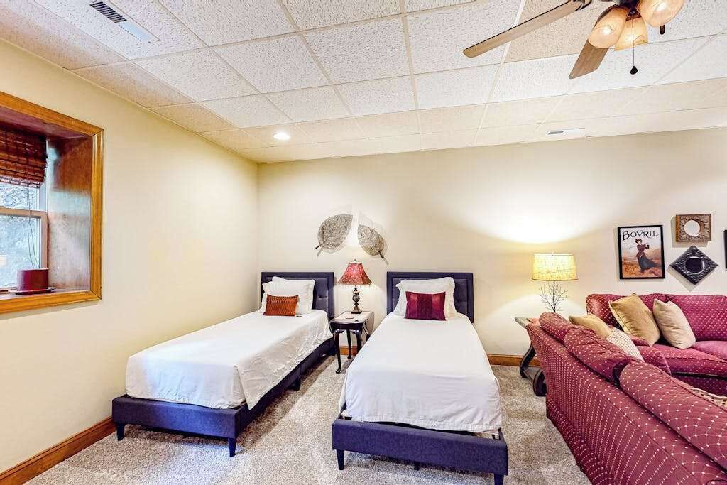 A hotel room with two beds, a red sofa, a ceiling fan, and a window with blinds.