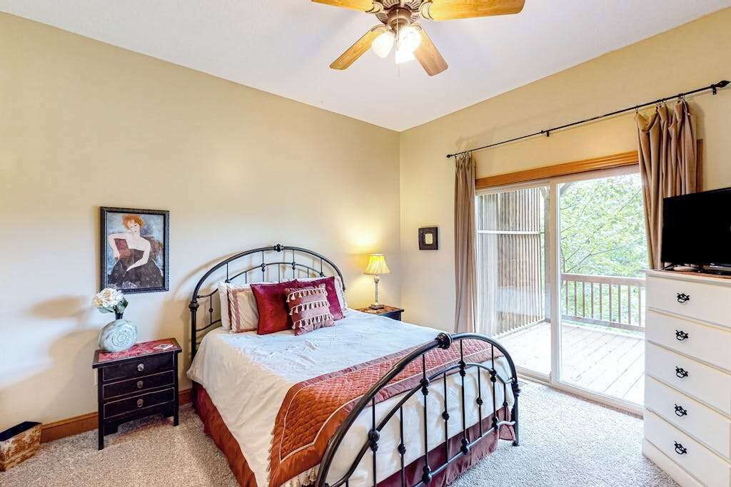 Une chambre à coucher comprenant un lit, une table de chevet, un téléviseur, un ventilateur de plafond et un balcon avec des rideaux.